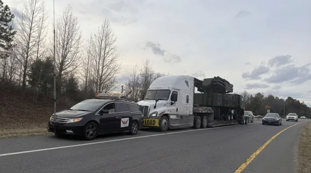 Black TCB Escorts pilot car stopped beside a truck hauling a military vehicle load on a country road