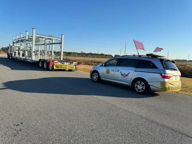 Silver TCB Escorts pilot car with red flags following a trailer hauling industrial equipment
